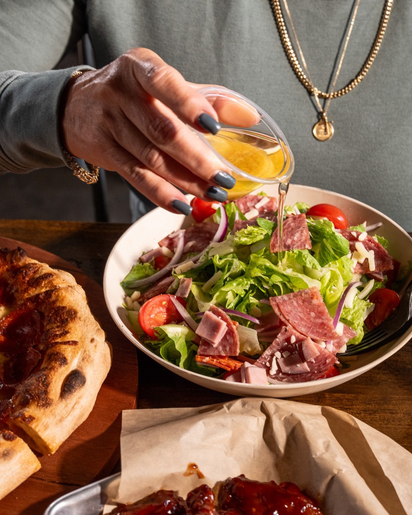 hand pouring italian dressing salad with fresh tomatoes, sliced ham, salami, and onions