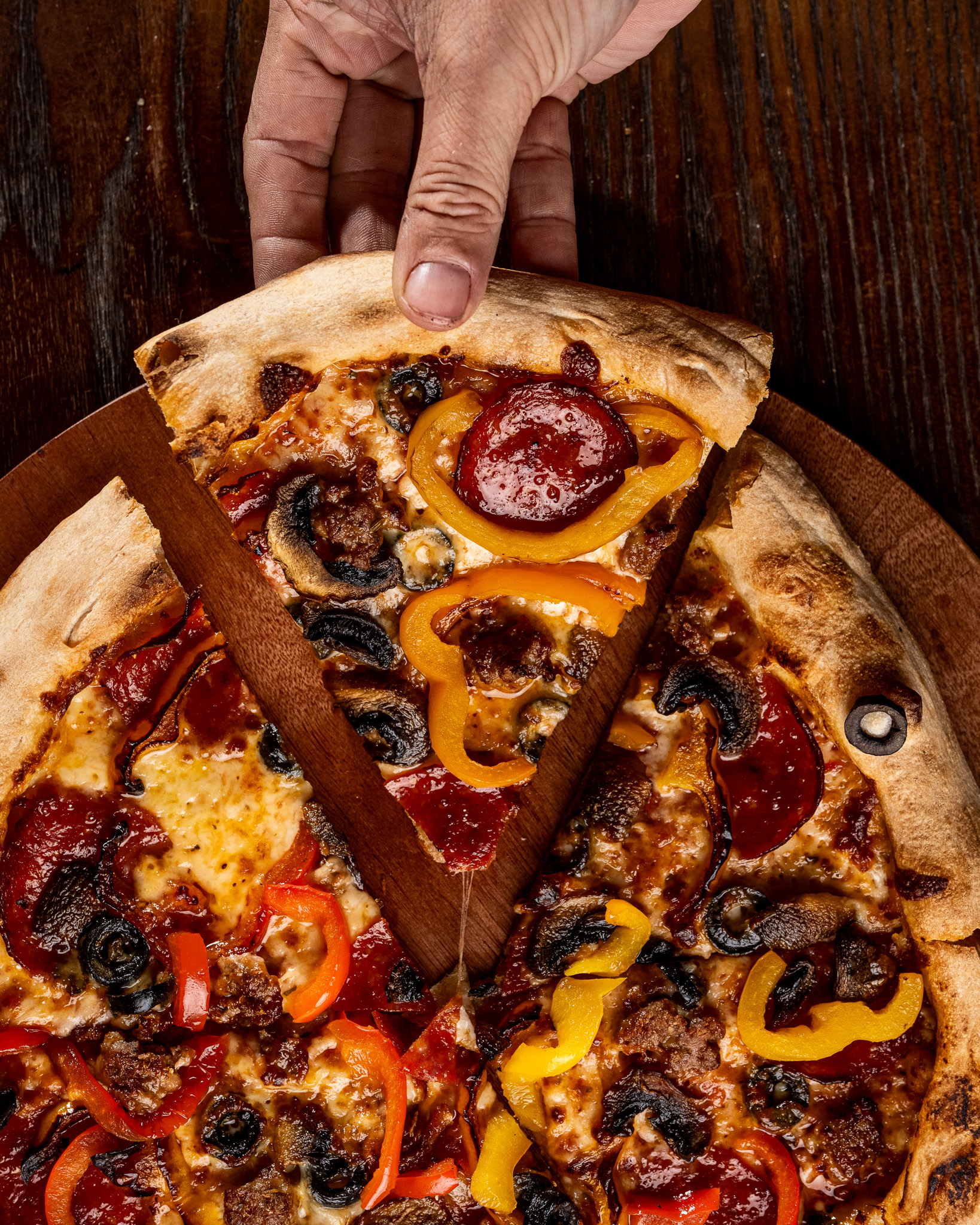 hand grabbing a slice of pizza with roasted pepperoni, mushrooms, sausage, and bell peppers